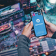 A person holding a phone with the OpenSea logo stands far above people walking on a street below.